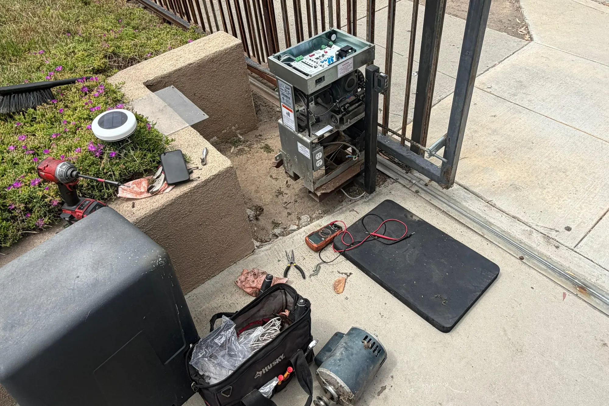 A gate operator dismantled on site with hand tools, a drill and a removed motor laid out on the driveway